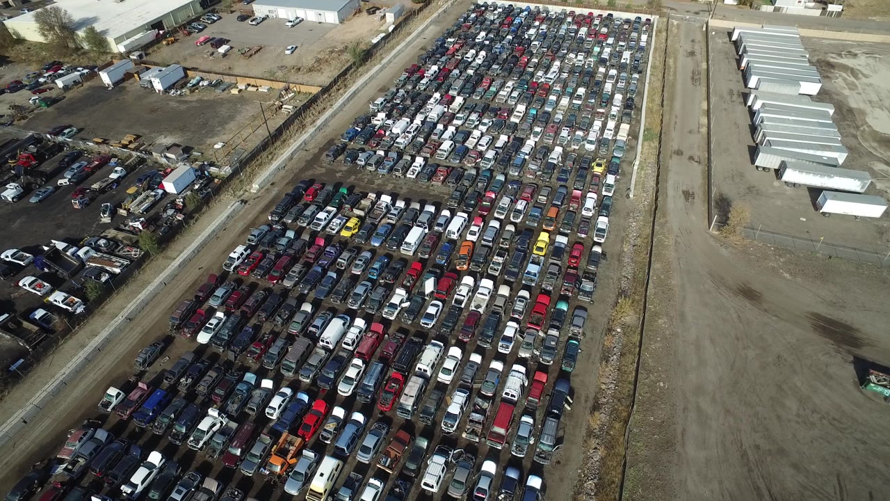 High aerial view in Westminster, Colorado, showing tightly parked cars and trucks, pale storage-like buildings on one side, dirt ground, and no visible people.