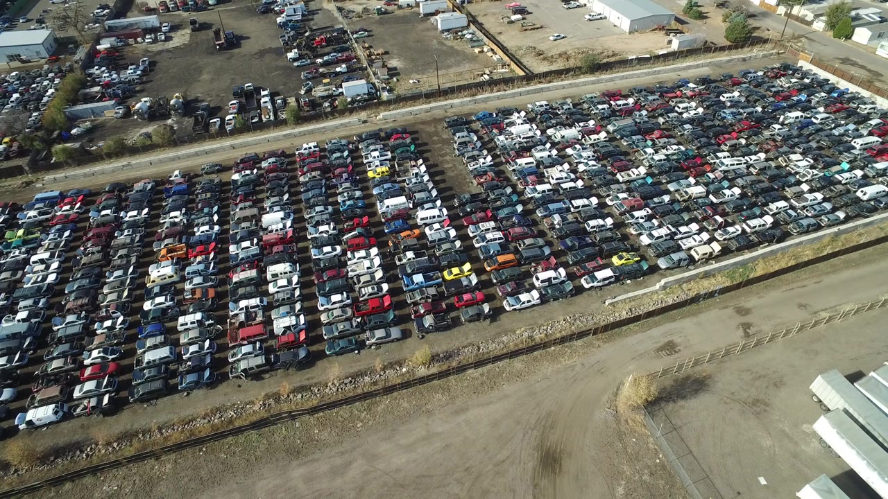 Aerial frame in Westminster, Colorado showing tightly packed rows of cars in a large industrial lot, a rail line along the lower edge, warehouse buildings beyond, and no visible people.