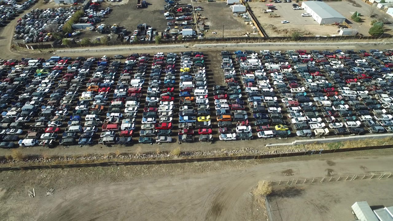 High aerial frame in Westminster, Colorado with packed car rows across the lower half, a dirt road through the center, and pale-roofed buildings beyond, with no visible people discernible.
