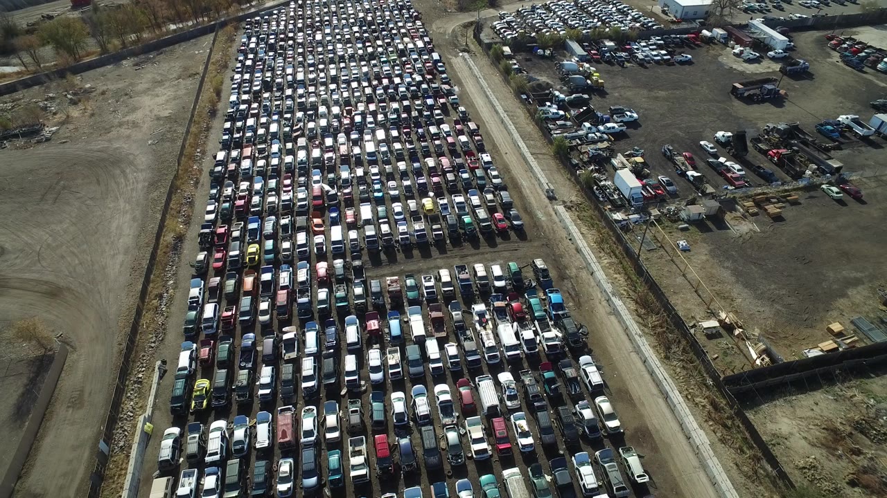 High oblique view in Westminster, Colorado, looking across tightly parked cars, a pale service road, open dirt areas, and no visible people.
