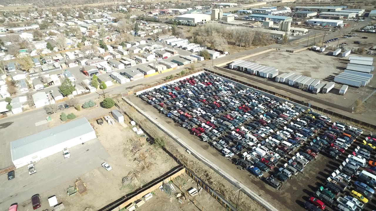 Aerial view over a crowded vehicle lot beside industrial buildings and suburban houses.
