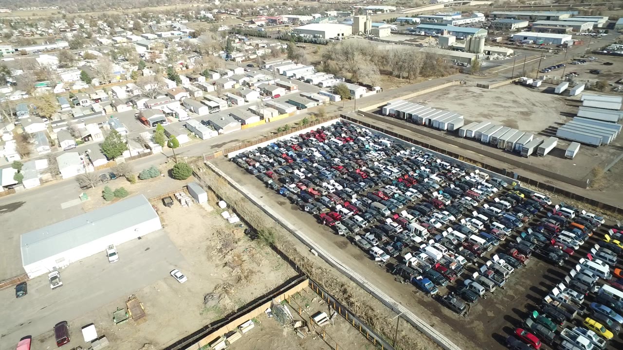Aerial frame in Westminster, Colorado with the packed car lot centered, warehouses and storage-style buildings behind it, and no visible people.