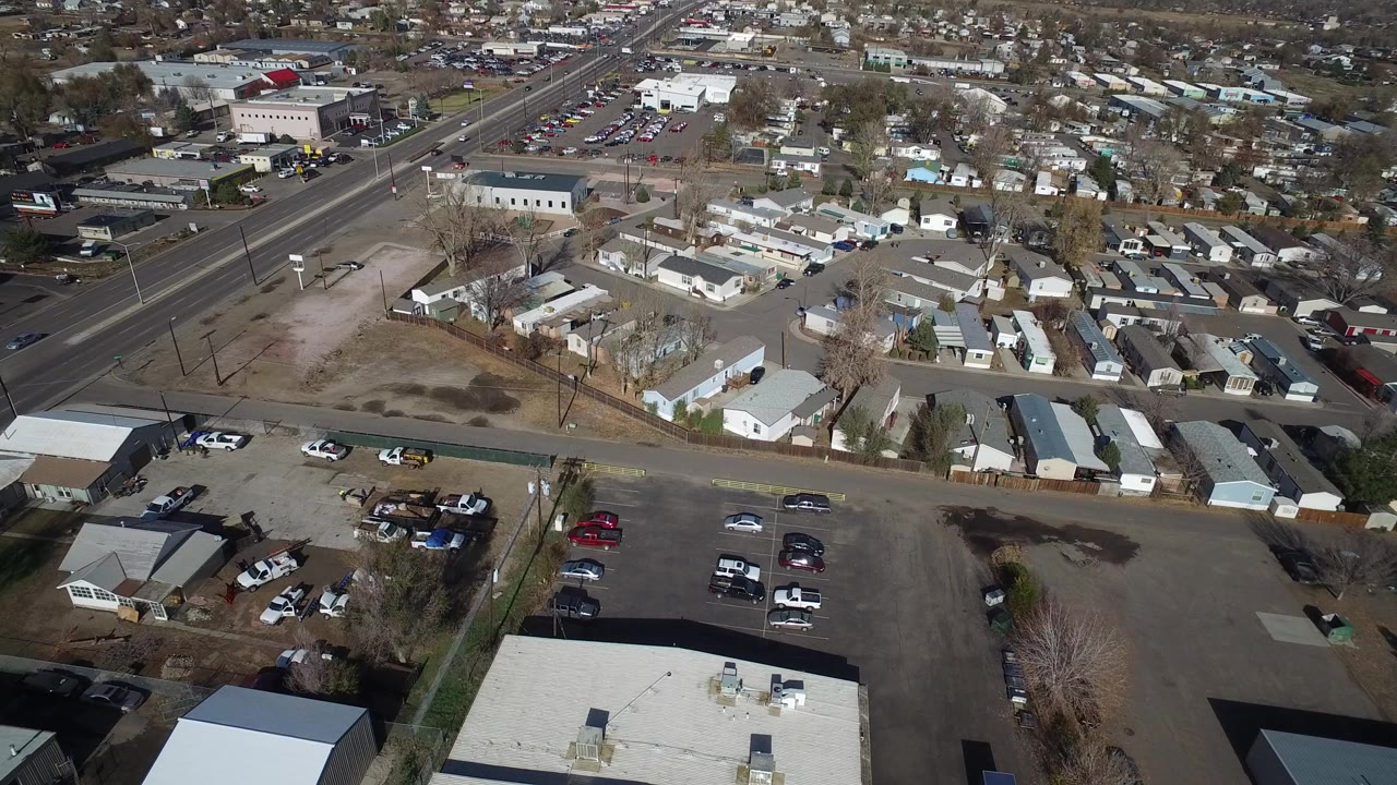 Opening aerial view in Westminster, Colorado, with a broad road crossing the upper left, parked cars in foreground lots, low buildings, small homes, bare trees, and no visible people.