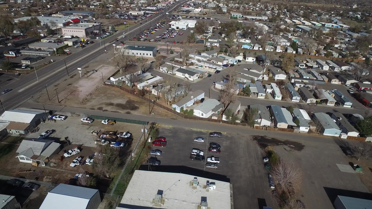 Aerial frame over Westminster, Colorado, showing foreground rooftops and parked vehicles, a diagonal arterial road, clustered small homes, light industrial buildings, and no visible people.