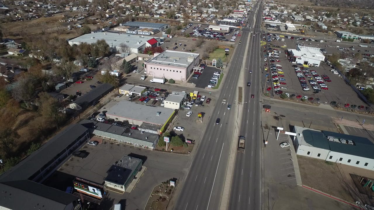 Opening aerial frame over Westminster, Colorado shows a multi-lane road bordered by businesses, parked cars, bare trees, and distant housing, with no visible people.