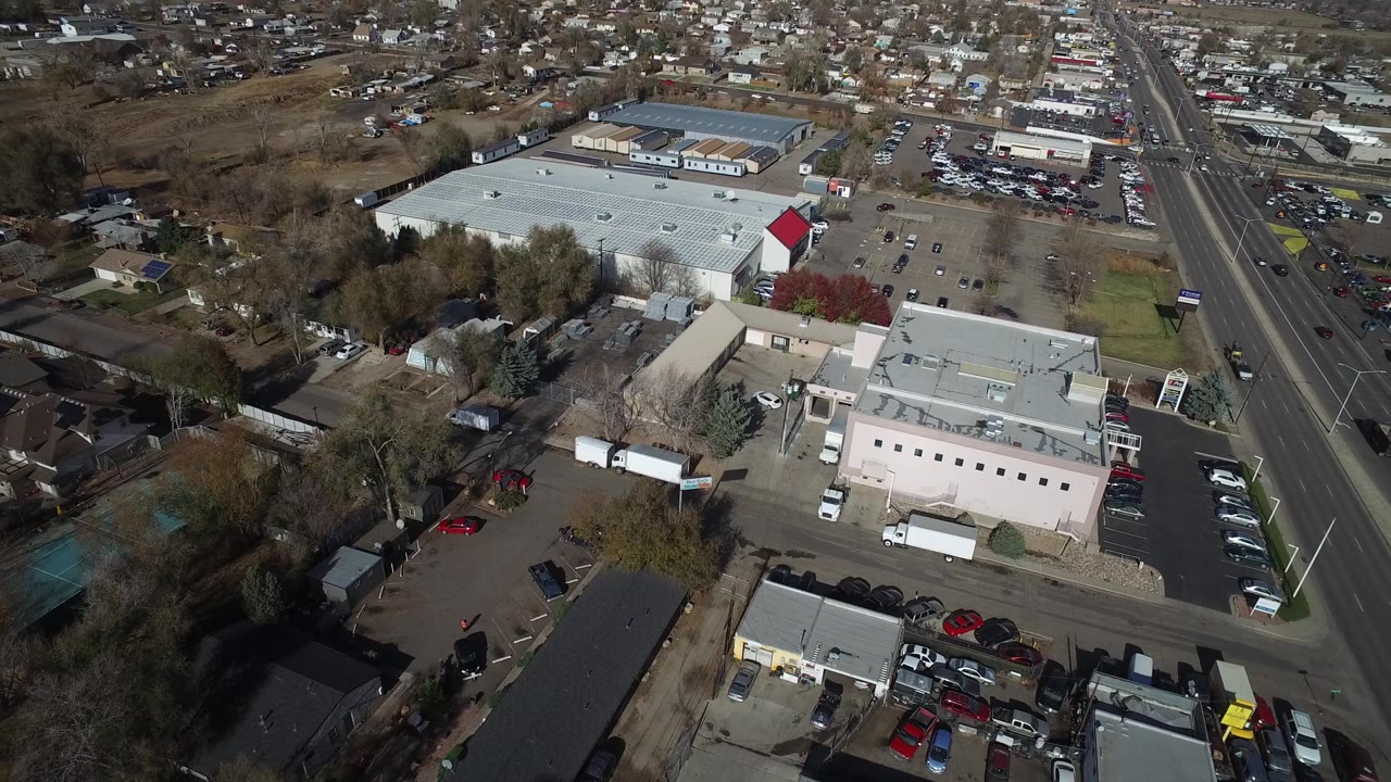 Westminster, Colorado aerial frame with flat-roof commercial buildings in the foreground, a wide road to the right, parked vehicles, trees, and distant houses, with no visible people.