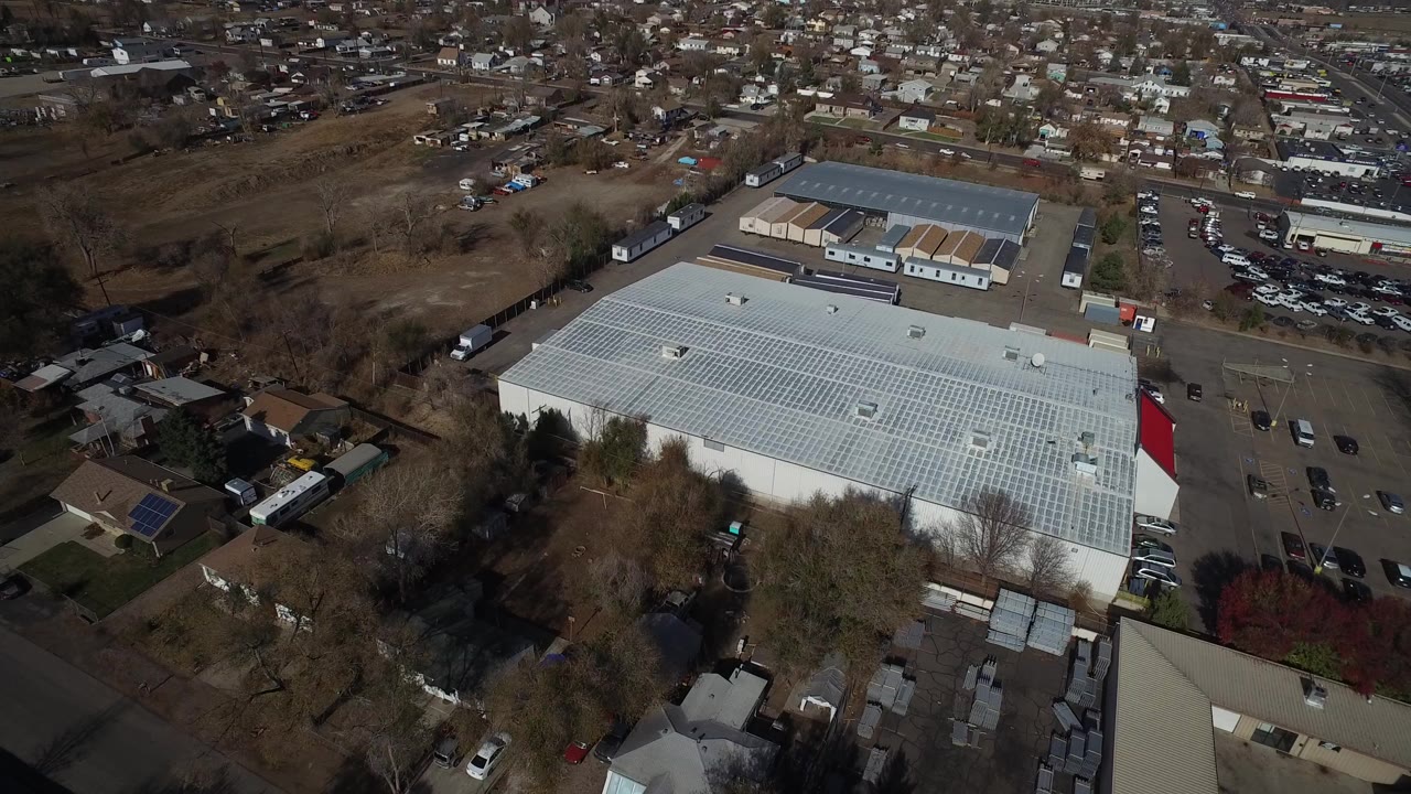 High aerial view in Westminster, Colorado showing a large pale-roofed commercial building, parked vehicles, nearby houses, bare trees, and no visible people.