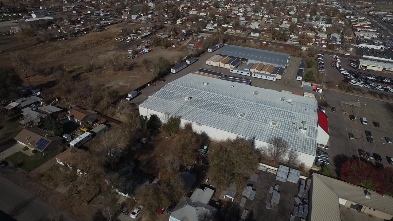 Aerial frame in Westminster, Colorado with the pale commercial roof centered, a road and parked cars to the right, residential blocks beyond, and no visible people.