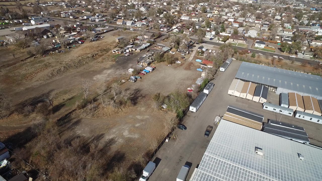 Oblique aerial frame in Westminster, Colorado with a large gray roof at bottom right, a paved access road, dry lots, scattered trees, parked cars, and houses beyond with no visible people.