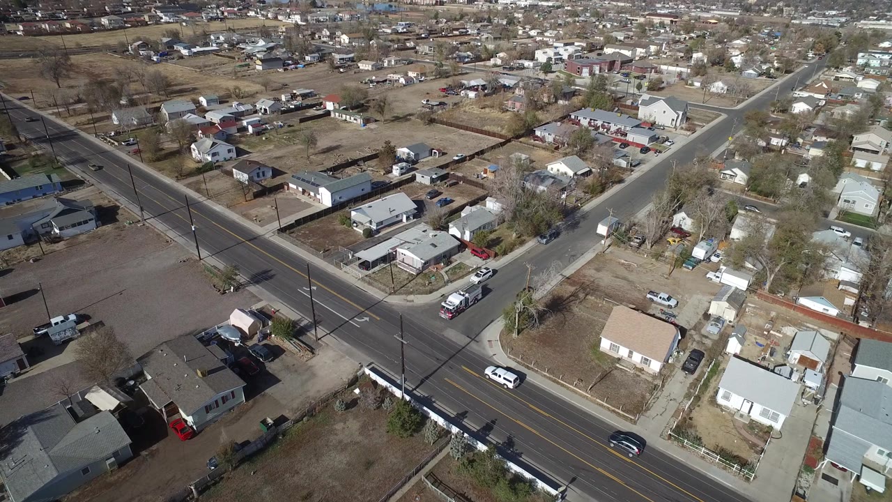 Aerial frame over Westminster, Colorado, showing the main road cutting diagonally past side streets, dry yards, low homes, parked vehicles, and no visible people.