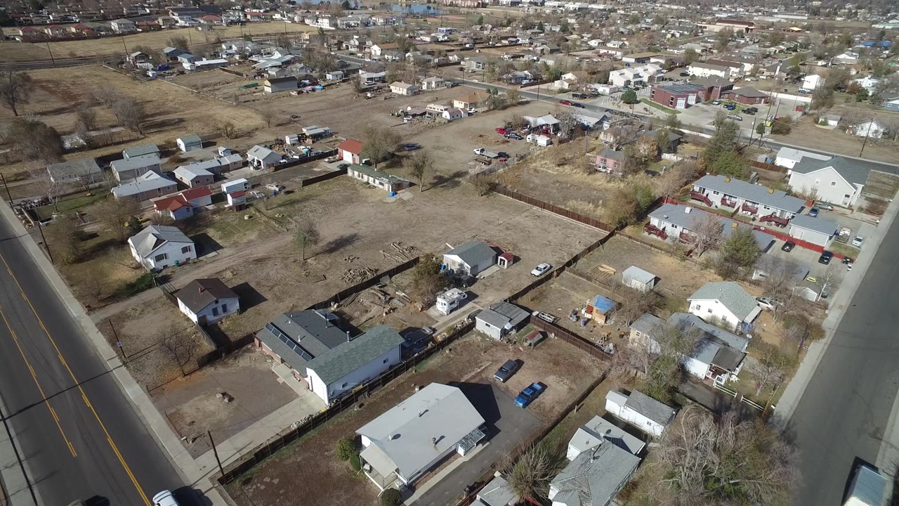 Aerial frame in Westminster, Colorado with a curved paved road on the right, low houses, sheds, parked vehicles, brown vegetation, and no visible people.
