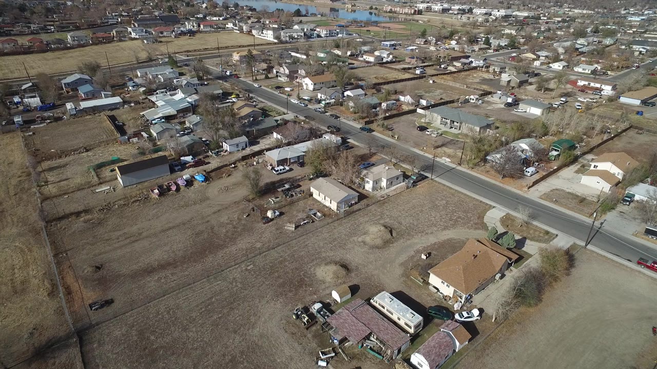Aerial frame in Westminster, Colorado showing a diagonal road, scattered single-story homes, parked cars, sheds, open brown yards, and no visible people.