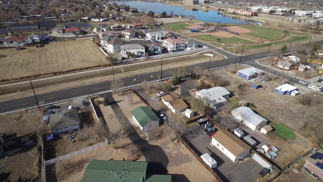 Opening aerial frame over Westminster, Colorado homes with a multi-lane road, parked cars, brown lawns, ball fields, a lake in the distance, and no visible people.