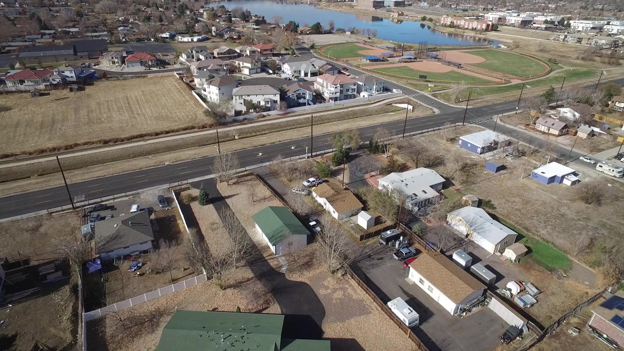 Aerial view in Westminster, Colorado with rooftops and yards below, a broad road crossing the frame, baseball diamonds behind it, water at the top edge, and no visible people.