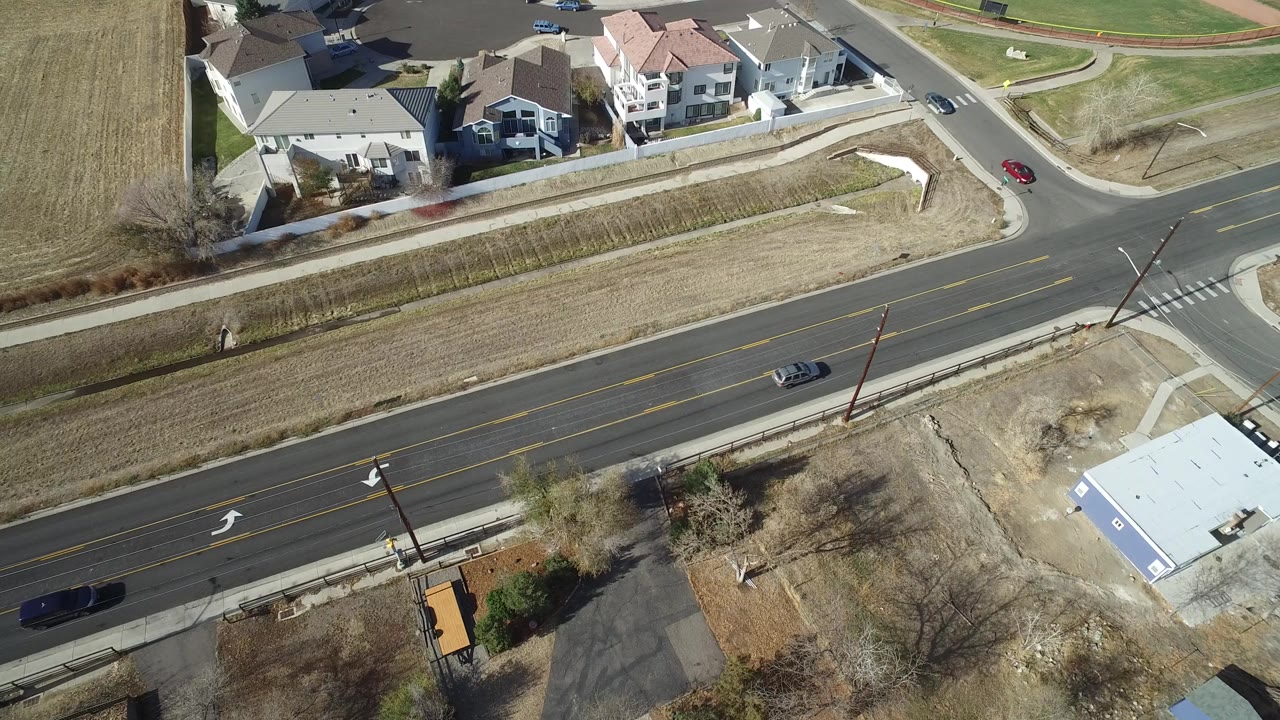 Aerial frame over Westminster, Colorado with a dark car on the main road, houses along the upper edge, sidewalks, open tan grass, and no visible people.