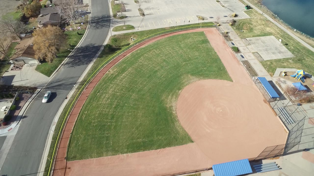 Overhead Westminster, Colorado frame with the green outfield centered, tan dirt infield to the right, red path curving along the edge, a road, parking spaces, water, and no visible people.