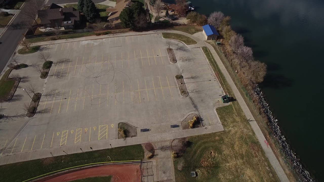 The second Westminster aerial frame holds over the empty lot and curved shoreline, with green-grey water on the right, houses beyond the lot, and no visible people.