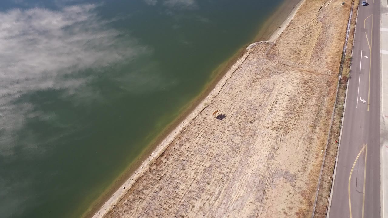 Aerial view of water, dry shoreline grass, and a paved road with a distant car.