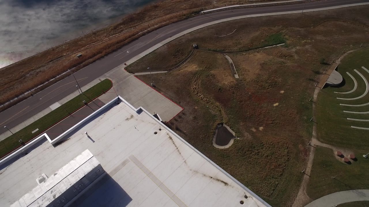 Westminster, Colorado aerial view with the roof edge lower left, a curving road near the top, a small pond, open grass, and curved concrete bands entering on the right, with no visible people.