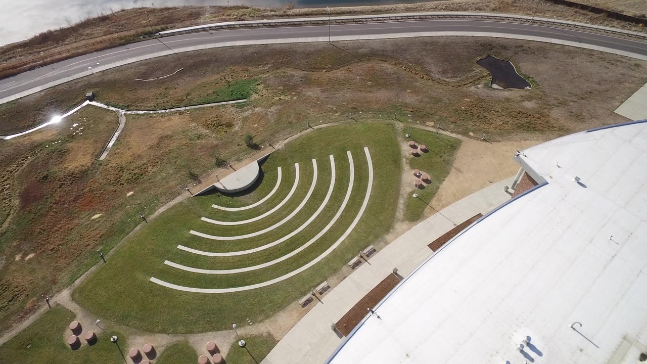 Overhead Westminster, Colorado frame with the amphitheater-like lawn more centered, a white roof at right, road at top, small ponds, paths, and no visible people.