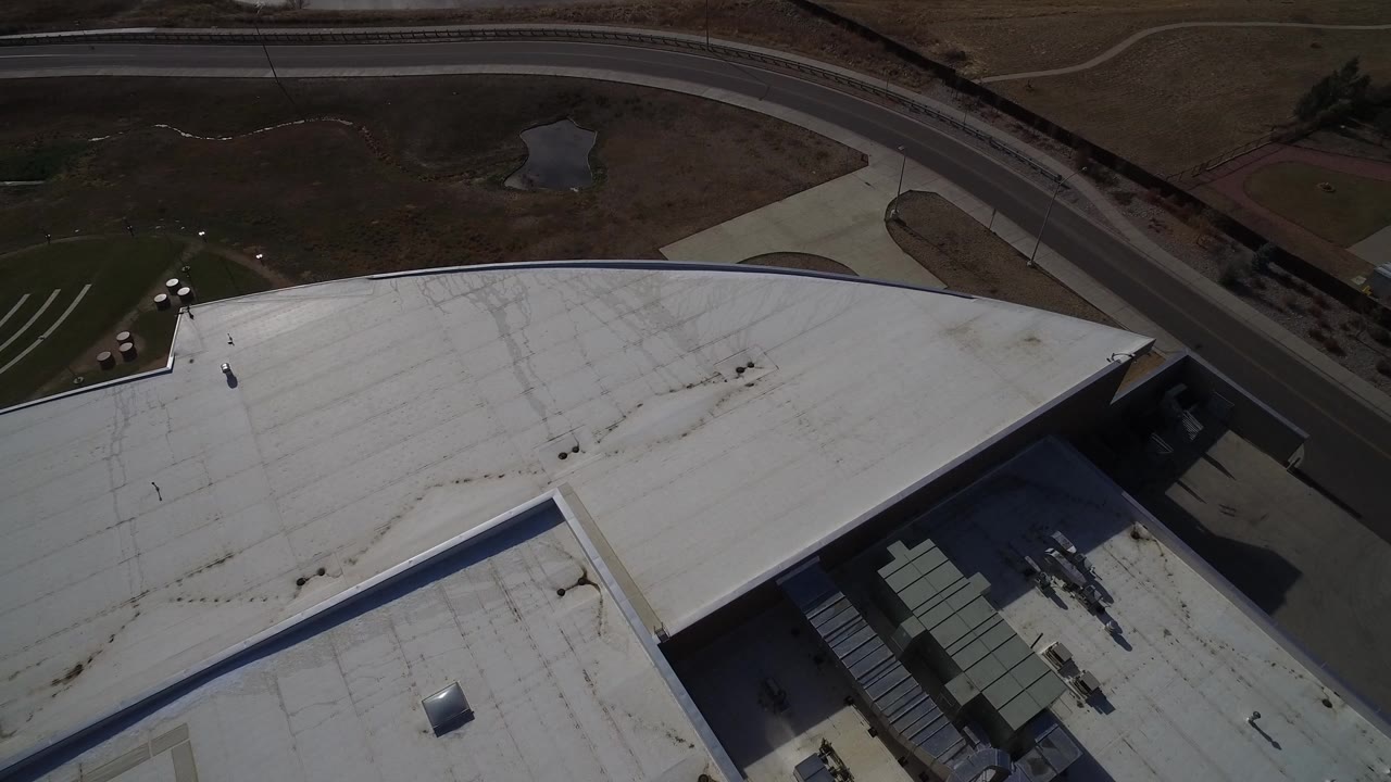 High aerial frame in Westminster, Colorado showing the broad white roof of an industrial building, a curved road above it, dry ground, and no visible people.