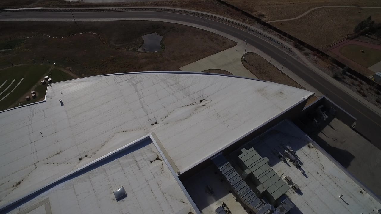Aerial view in Westminster, Colorado with the white-roofed factory dominating the lower frame, the road across the top, roof stains, and no visible people.