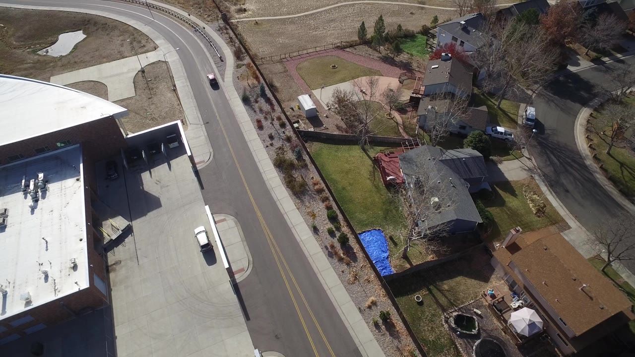 Aerial frame over Westminster, Colorado showing a curved road, a large flat-roof building, nearby houses, lawns, bare trees, and no clearly visible people.