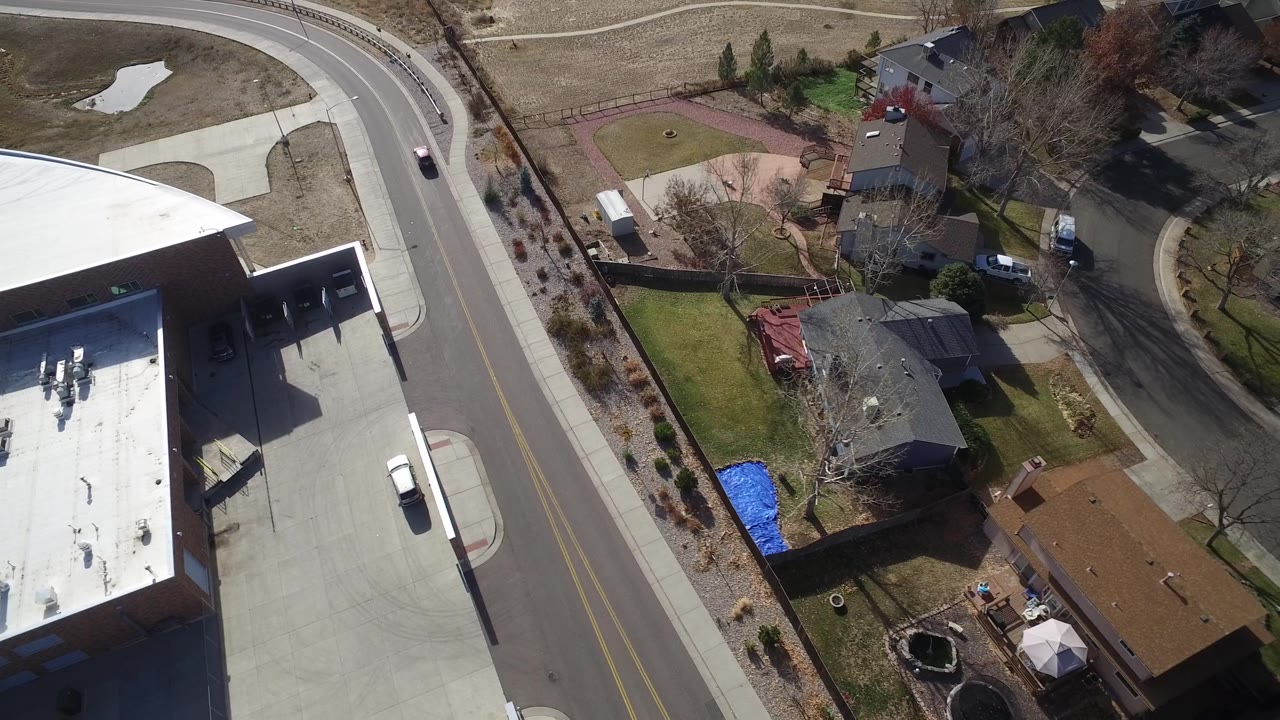 High-angle Westminster, Colorado view with the flat white roof at lower left, a two-lane residential road, detached homes, fenced yards, and no clearly visible people.