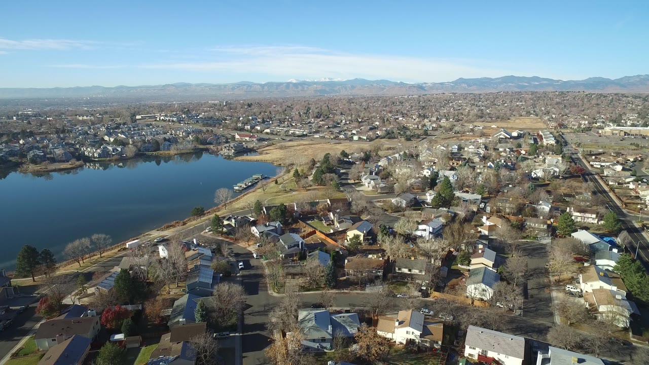 Westminster, Colorado neighborhood seen from above with the lake left of frame, rows of houses across the center, pale grass, distant mountains, and no visible people.