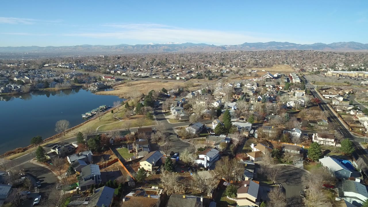 Aerial frame over Westminster, Colorado showing rooftops and bare trees in the foreground, a dark blue lake to the left, neighborhoods stretching back, and no visible people.