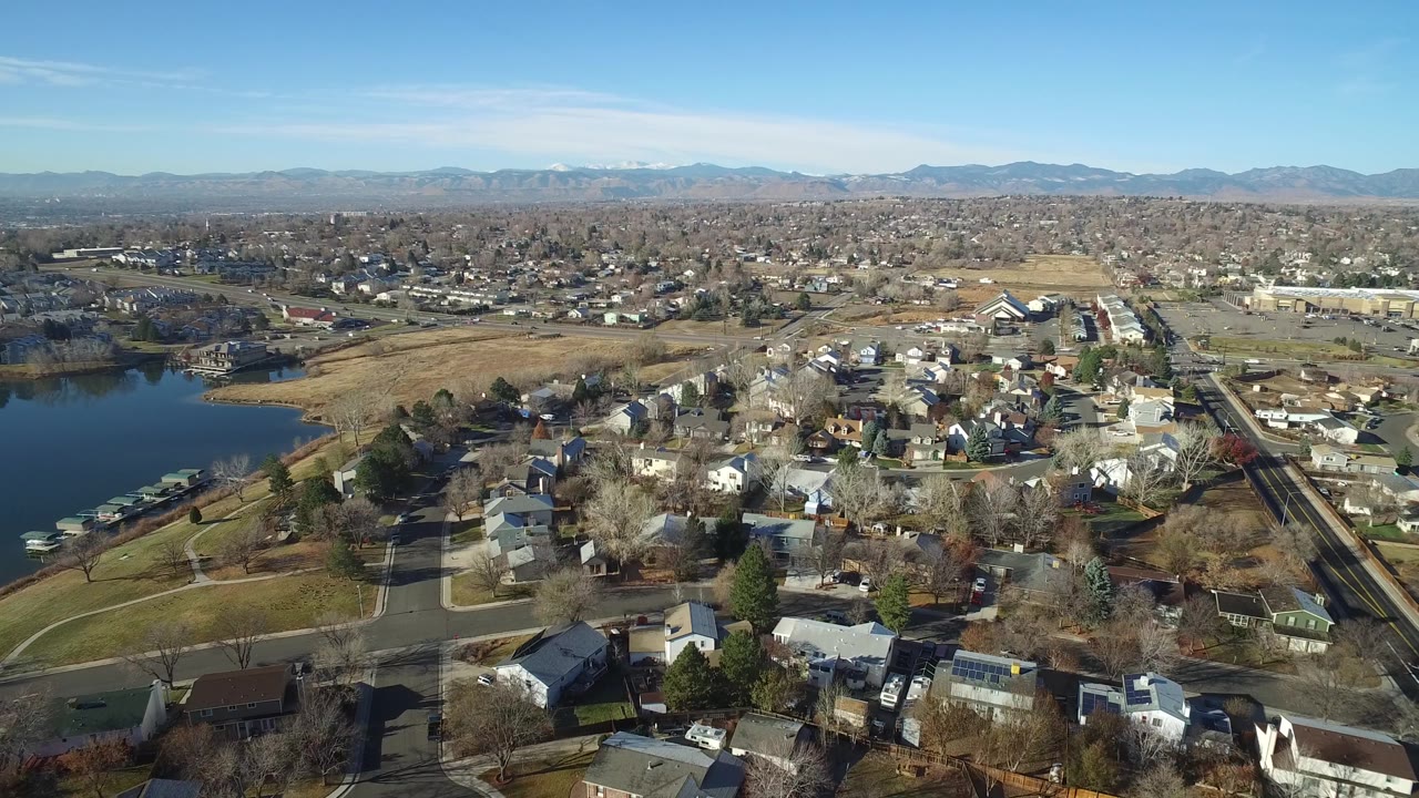 Aerial frame over Westminster, Colorado with the lake still visible at left, rows of private homes below, a road corridor to the right, tan fields in the middle distance, and no visible people.
