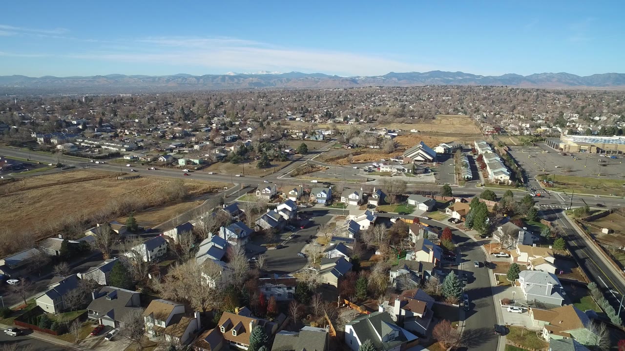 Opening aerial frame over Westminster, Colorado with residential rooftops in the foreground, open fields and roads in the midground, distant mountains on the horizon, and no visible people.