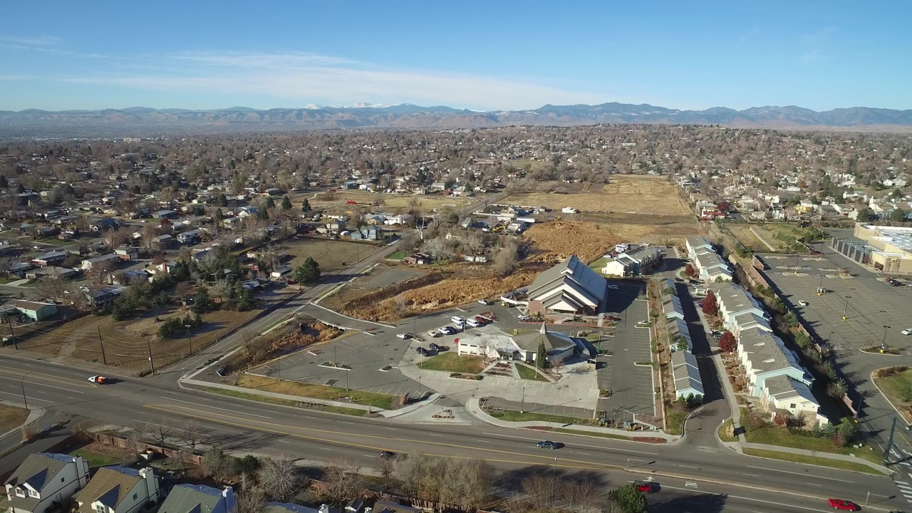 Aerial view in Westminster, Colorado with the main road crossing the foreground, low commercial structures and parking lots below, residential blocks beyond, distant mountains, and no visible people.