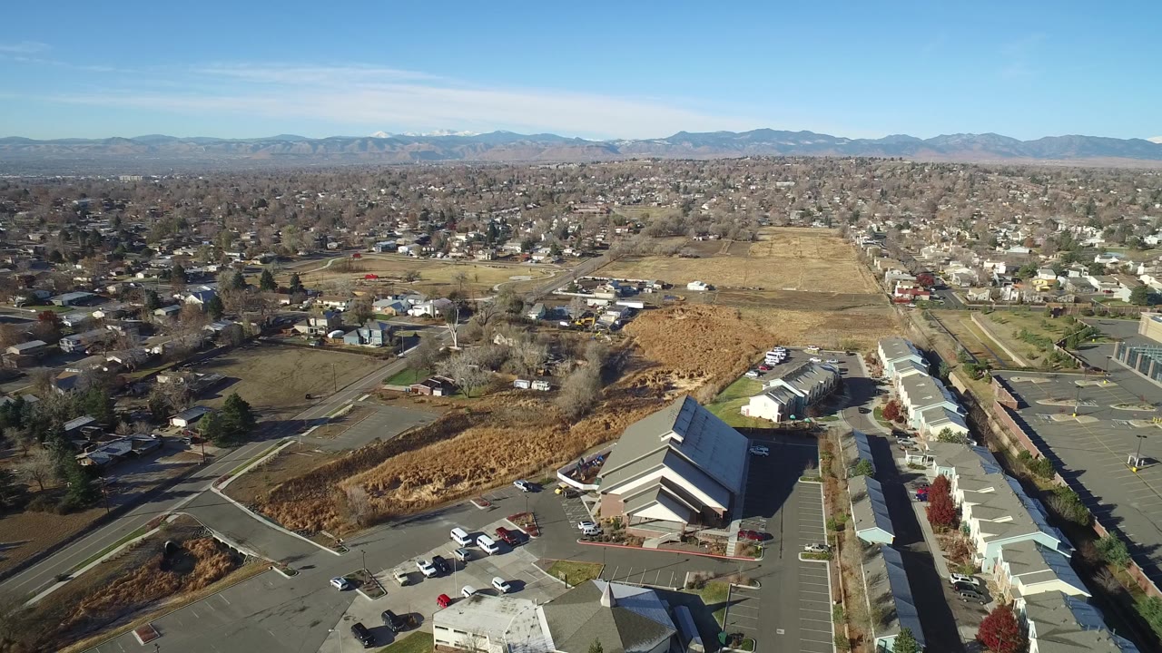Aerial view in Westminster, Colorado, with the foreground building lower in frame, dry tan fields through the center, neighborhoods beyond, mountains on the horizon, and no visible people.