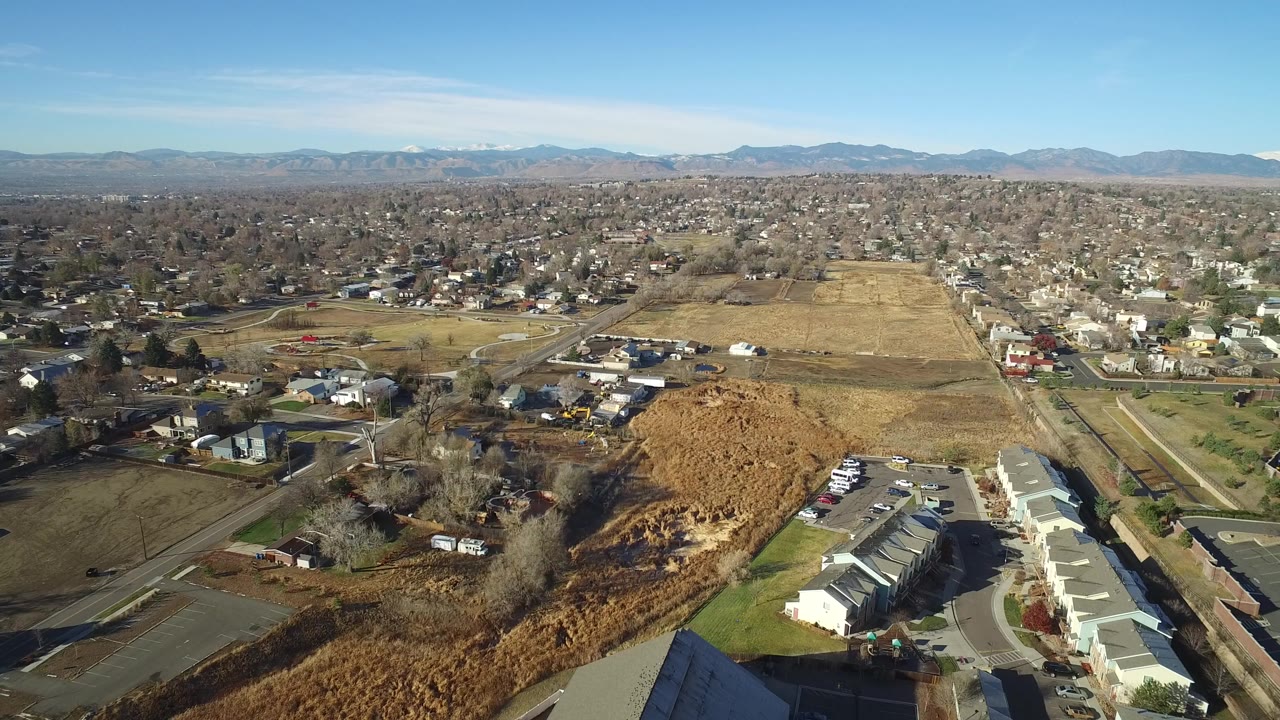 Wide aerial view over Westminster, Colorado with rooftops and parked cars in the foreground, open brown fields through the center, distant residential blocks, and no visible people.