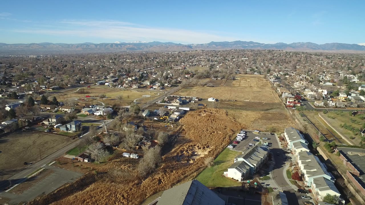 Westminster, Colorado aerial frame looking across a housing area beside dry open fields, with curving roads, tree-lined blocks, mountains on the horizon, and no visible people.