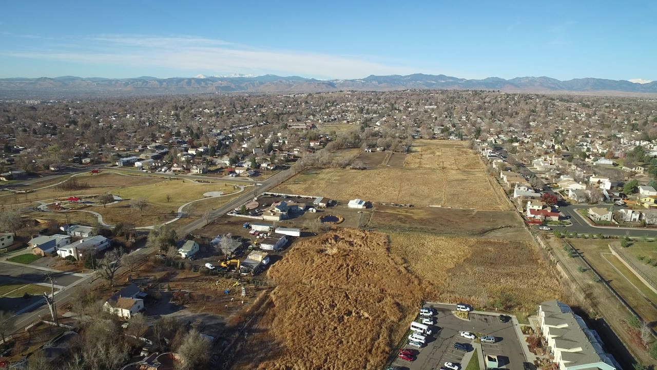 Aerial view over tan fields, suburban homes, roads, and distant mountains under blue sky.