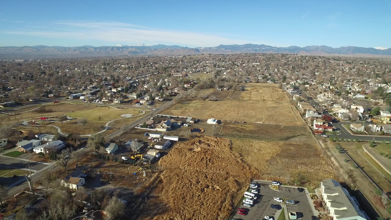 Second aerial frame over Westminster, Colorado showing a large rectangular tan field, nearby houses, curving roads, distant mountains, and no visible people.