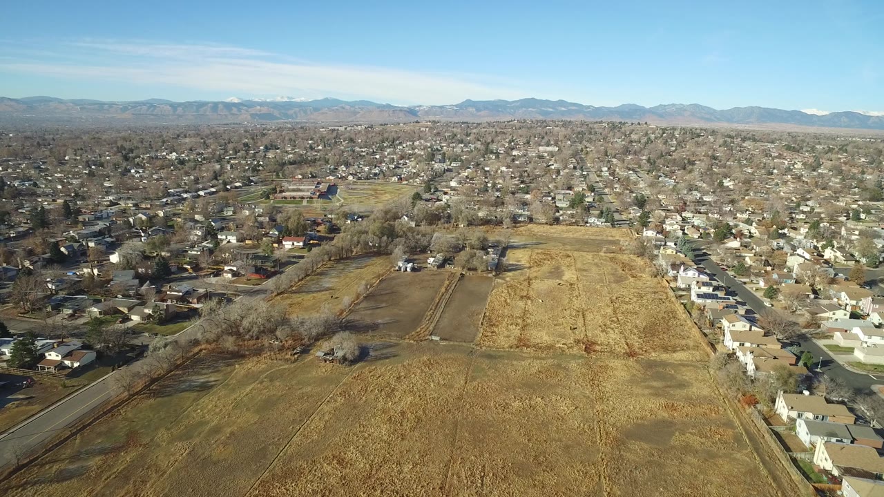Aerial view in Westminster, Colorado, with the dry field centered lower in frame, a row of pale homes to the right, dense suburbs beyond, and no visible people.