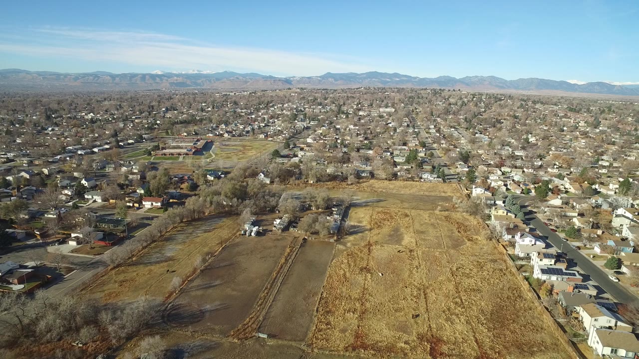 Aerial view in Westminster, Colorado, looking across brown and yellow open parcels toward gridded residential streets, low houses, mountains, and no visible people.