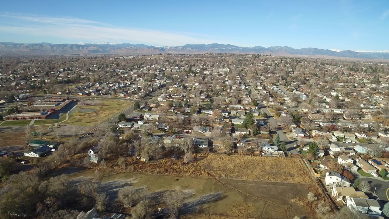 Aerial view of suburban houses, dry fields, trees, and distant mountains under blue sky.