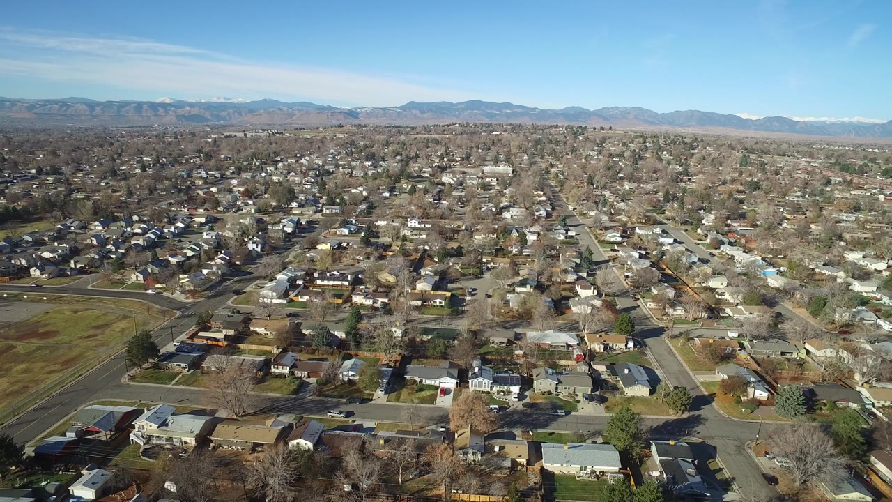Wide aerial view over Westminster, Colorado homes, streets, bare trees, and distant mountains under a clear blue sky, with no visible people.