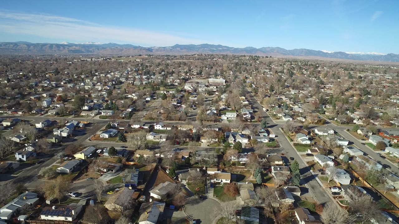 Wide aerial view over Westminster, Colorado residential blocks, with leafless trees, single-family homes, distant mountains, blue sky, and no visible people.
