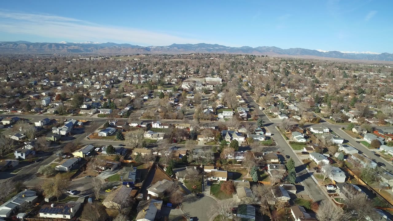 High view across Westminster, Colorado homes and crossing residential streets, with winter-bare trees, parked vehicles, foothills, and no visible people.