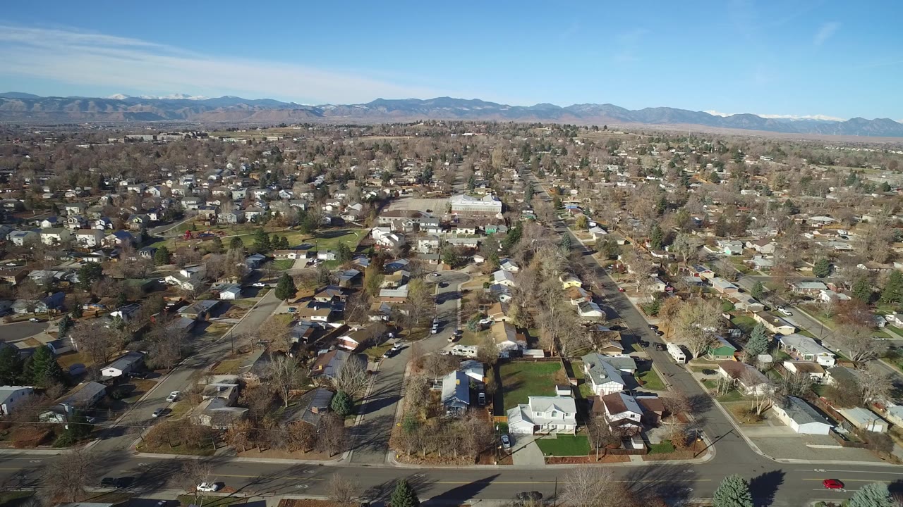 Second aerial frame over Westminster, Colorado with a central road corridor, clustered houses, bare trees, low winter color, distant mountains, and no visible people.