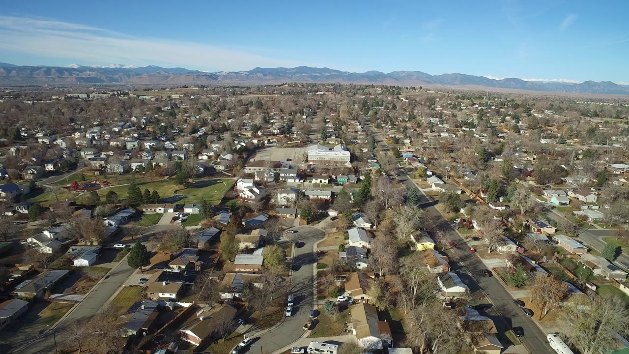 Aerial view of Westminster, Colorado homes, streets, bare trees, and distant mountains under a clear blue sky, with no visible people.