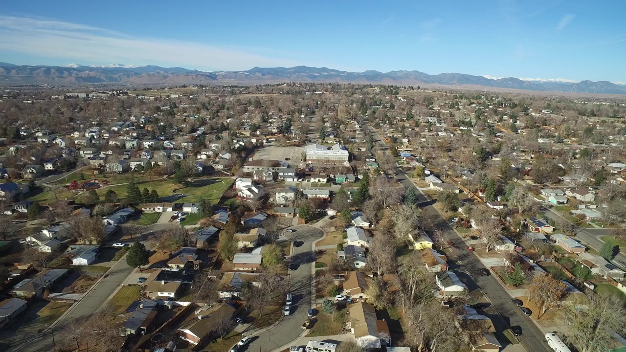 High aerial frame over Westminster, Colorado residential blocks, a larger light-roofed building, tree-lined streets, and distant mountains, with no visible people.