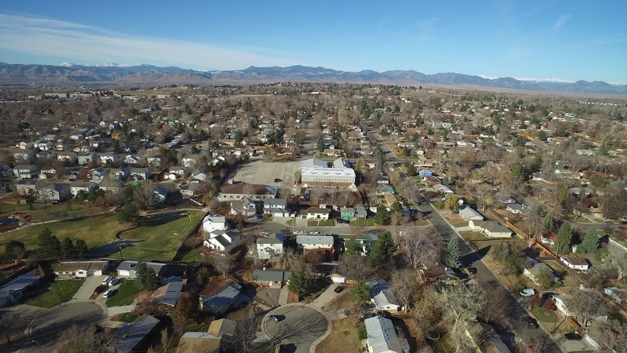 Wide aerial view over Westminster, Colorado homes, leafless trees, residential streets, a large light-roofed building, and distant mountains under clear sky, with no visible people.