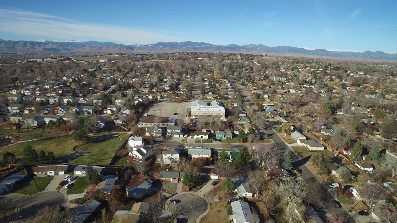 High aerial frame of Westminster, Colorado residential blocks with rooftops, bare trees, a straight road through the center-right, and mountains along the far horizon, with no visible people.