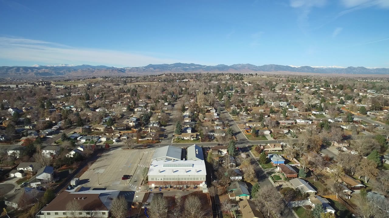 Opening aerial frame over Westminster, Colorado with a light-roofed building in the lower foreground, residential streets beyond, distant mountains, blue sky, and no visible people.